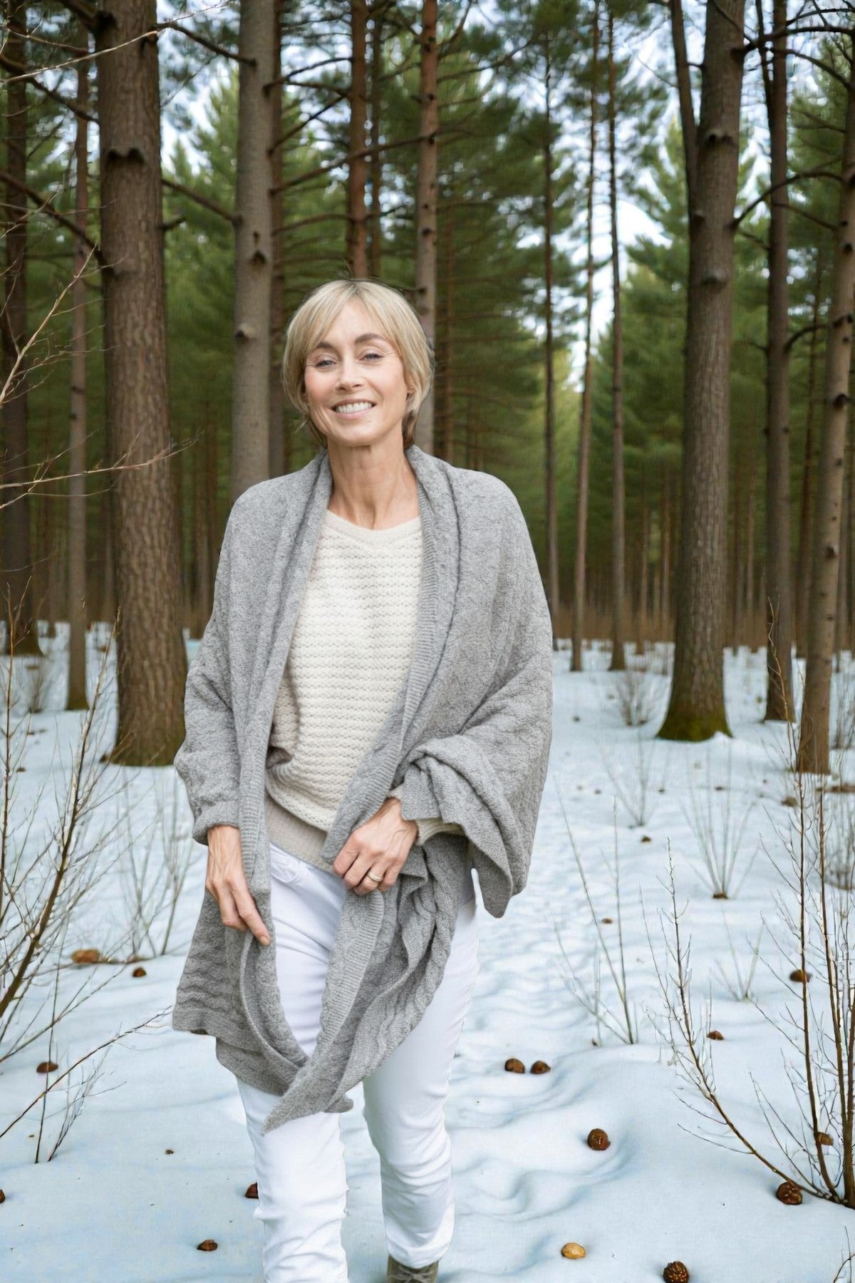 Woman in a gray cardigan and white pants standing in a snowy forest.